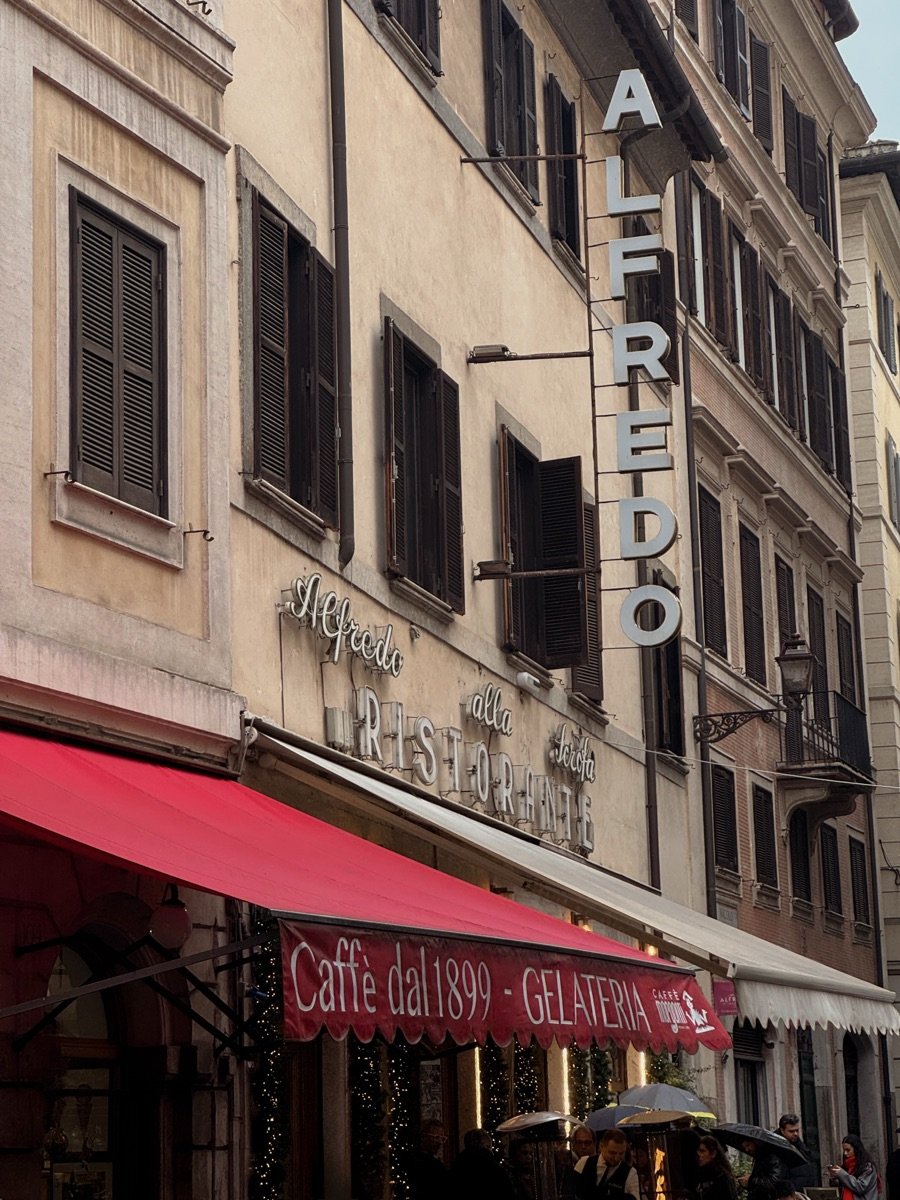 The corner of Alfredo alla Scrofa in Rome seen from the street, the restaurant&rsquo;s vertical sign above a red awning lettered Caff&egrave; dal 1899.