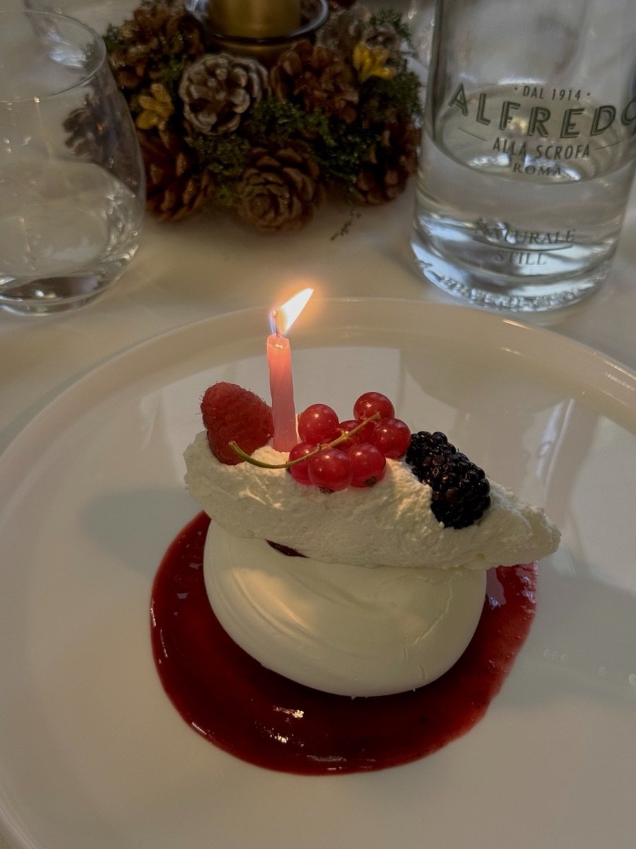 A meringue and cream dessert with raspberries, redcurrants and a blackberry on a pool of red fruit coulis, a lit candle in the centre.