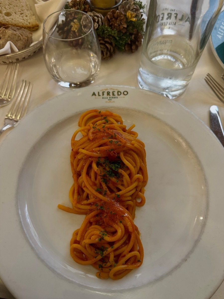 Spaghetti al pomodoro plated in a long curve on a white plate, bread and water on the linen behind it.