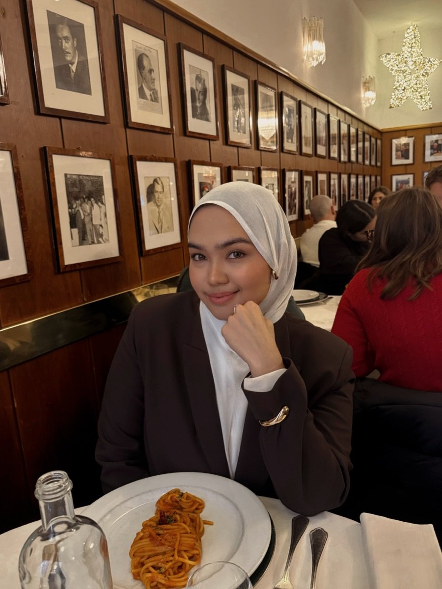 Riva seated in the dining room in a brown blazer and white headscarf, a wall of framed portraits behind her and a plate of pasta in front.