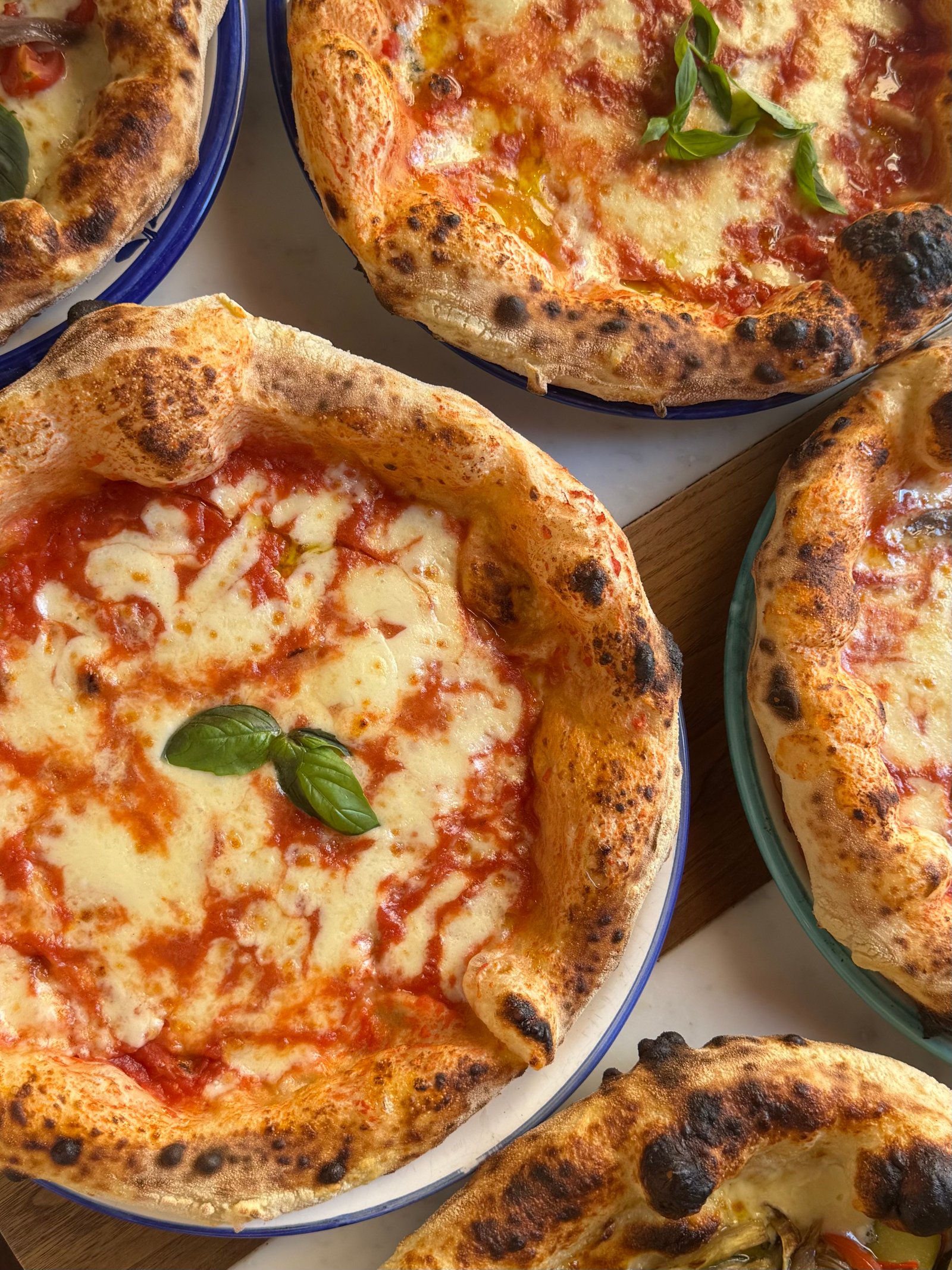 Five Neapolitan pizzas on enamel plates seen from directly above on a marble table, blistered crusts and basil.