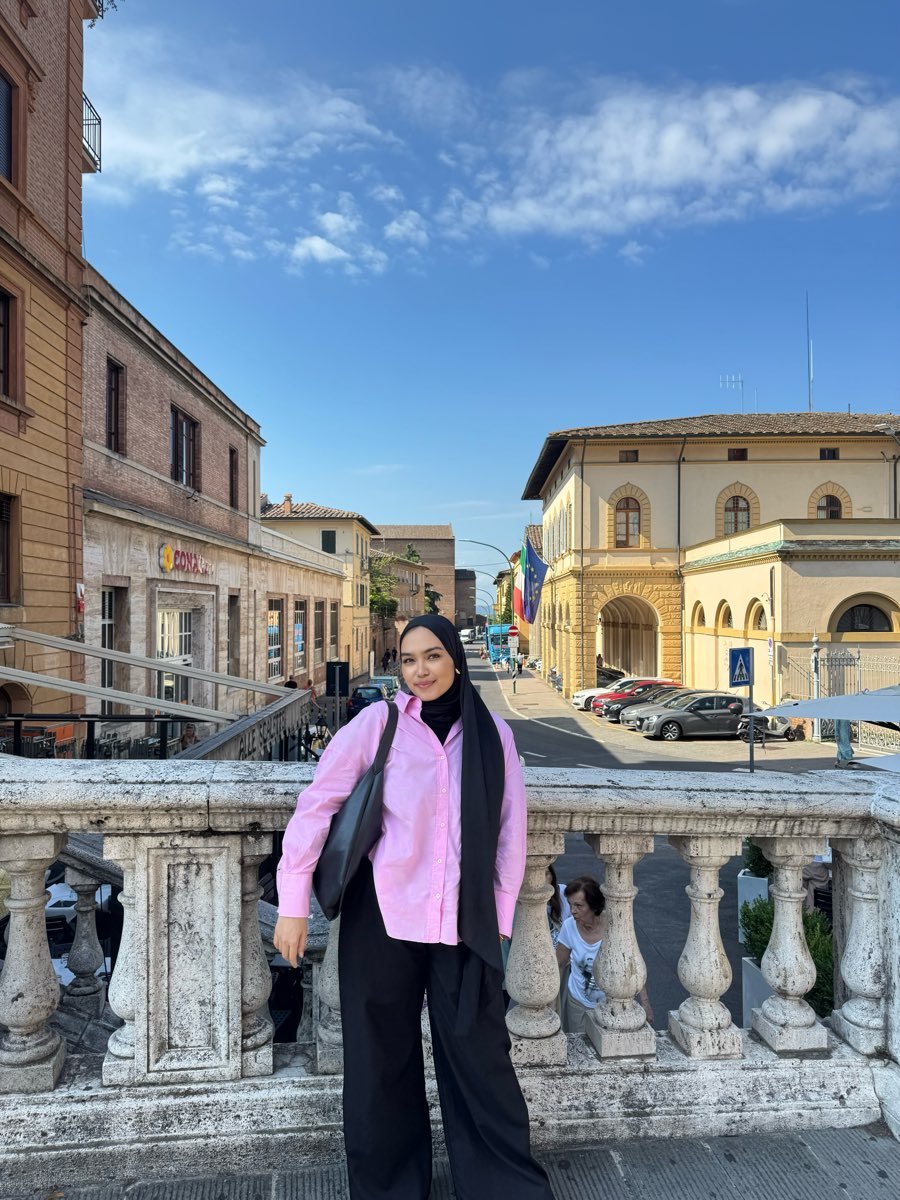 Riva in a pink shirt and black trousers standing at a stone balustrade above a street, buildings and flags behind her.
