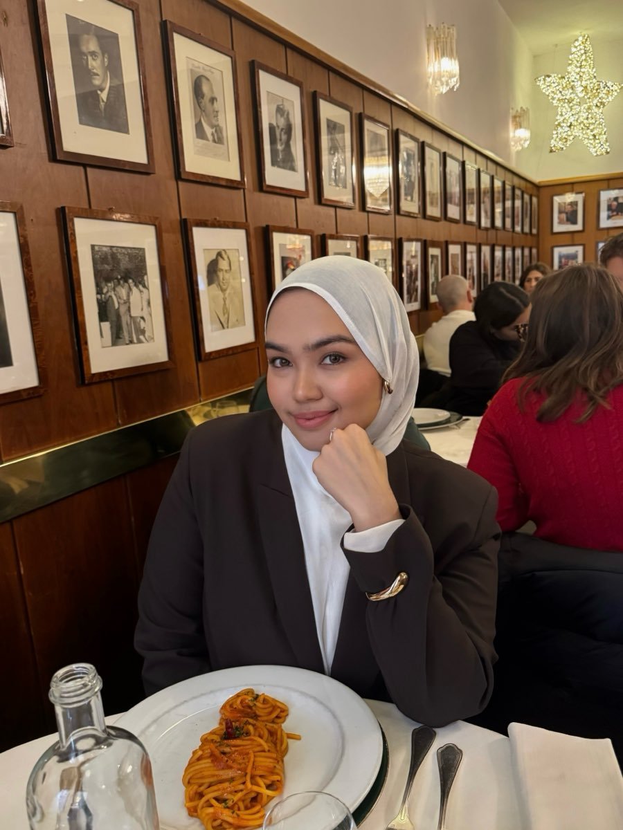 Riva seated at a restaurant table in a dark blazer, a wall of framed black and white photographs behind her and a plate of pasta in front of her.