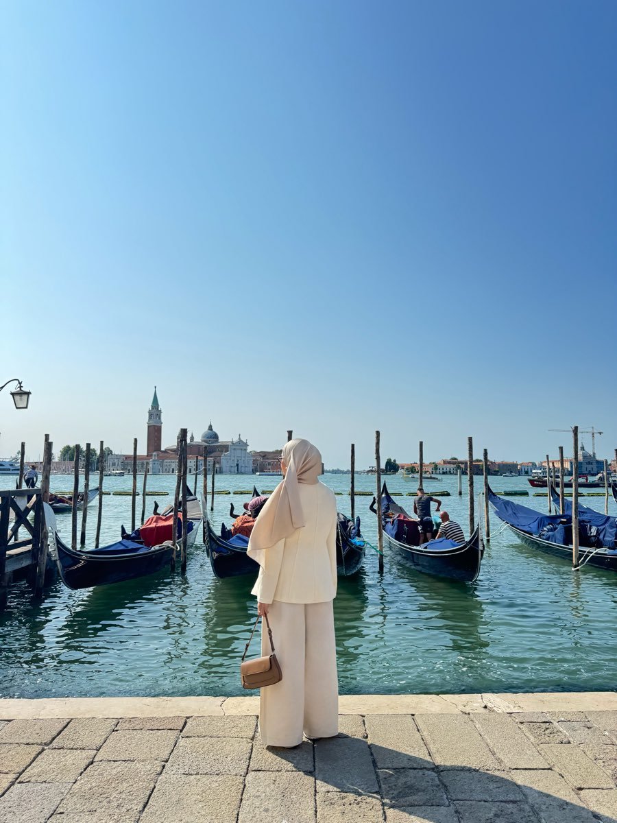 Riva standing on the waterfront in Venice, seen from behind, looking out across the water towards a domed church with moored boats in front of her.