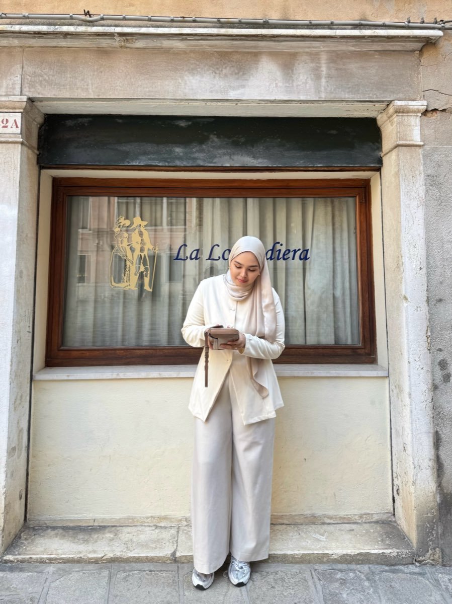 Riva in a cream suit standing against the stone frame of an old shopfront window, looking down at something in her hands.