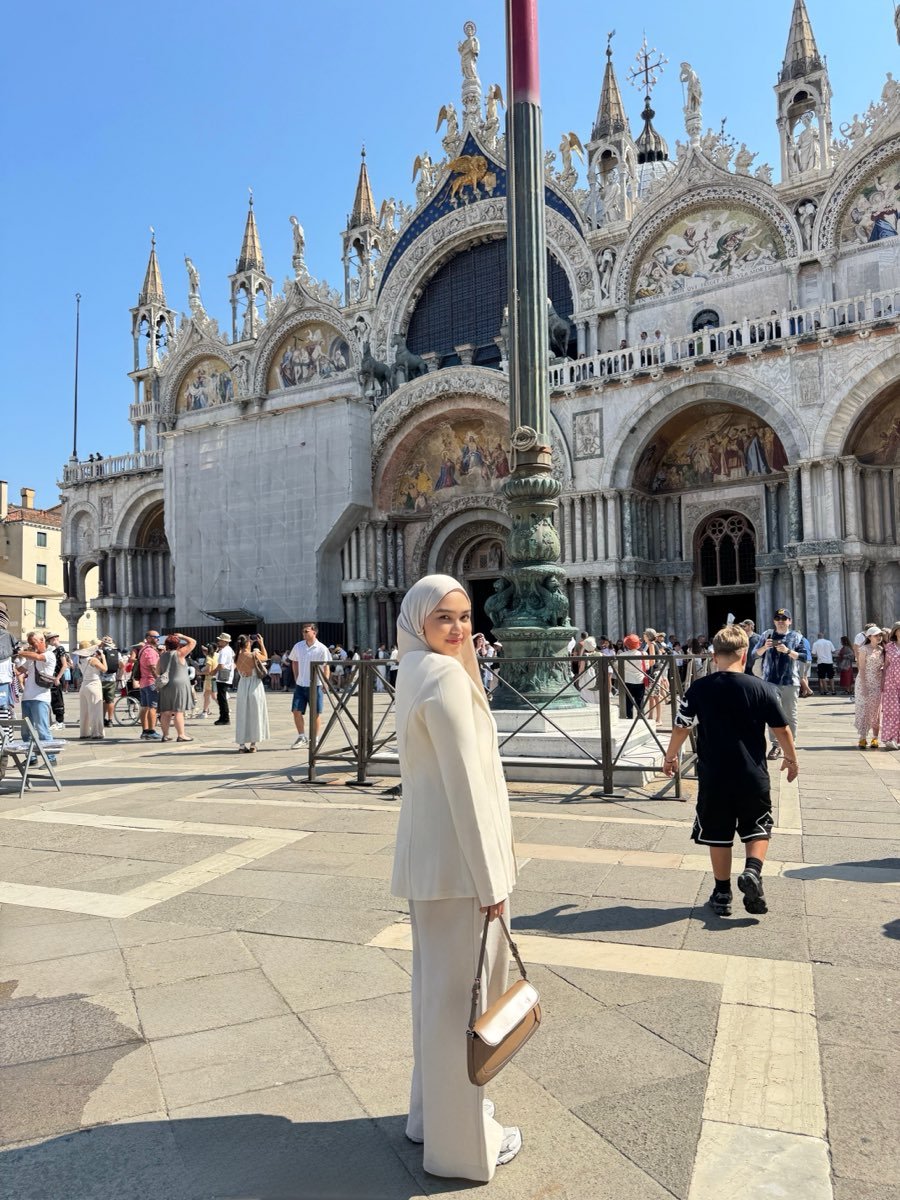 Riva in a cream suit standing in a square in front of the domed and arched facade of a basilica.